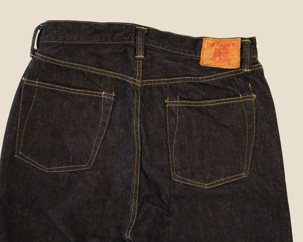 Close-up back view of dark indigo denim jeans showing the waistband, belt loops, two rear patch pockets, contrast yellow stitching, and a tan leather brand patch on a light beige background.