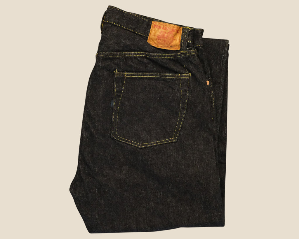 A folded pair of dark indigo denim jeans showing the back pocket and waistband patch, photographed against a solid light beige background.