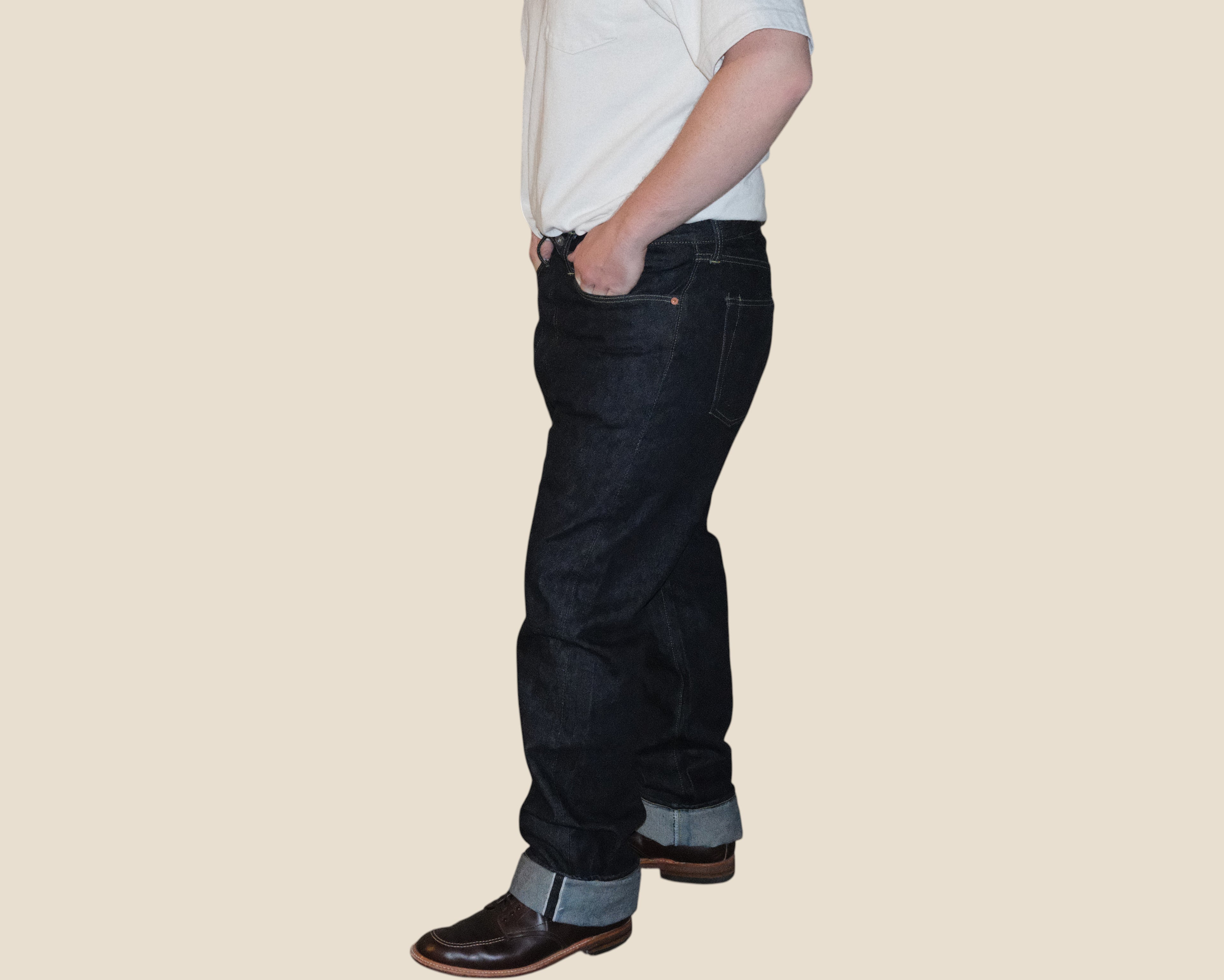 A side profile of a person wearing dark blue denim jeans with rolled cuffs, paired with a white shirt and brown shoes, against a solid beige background.