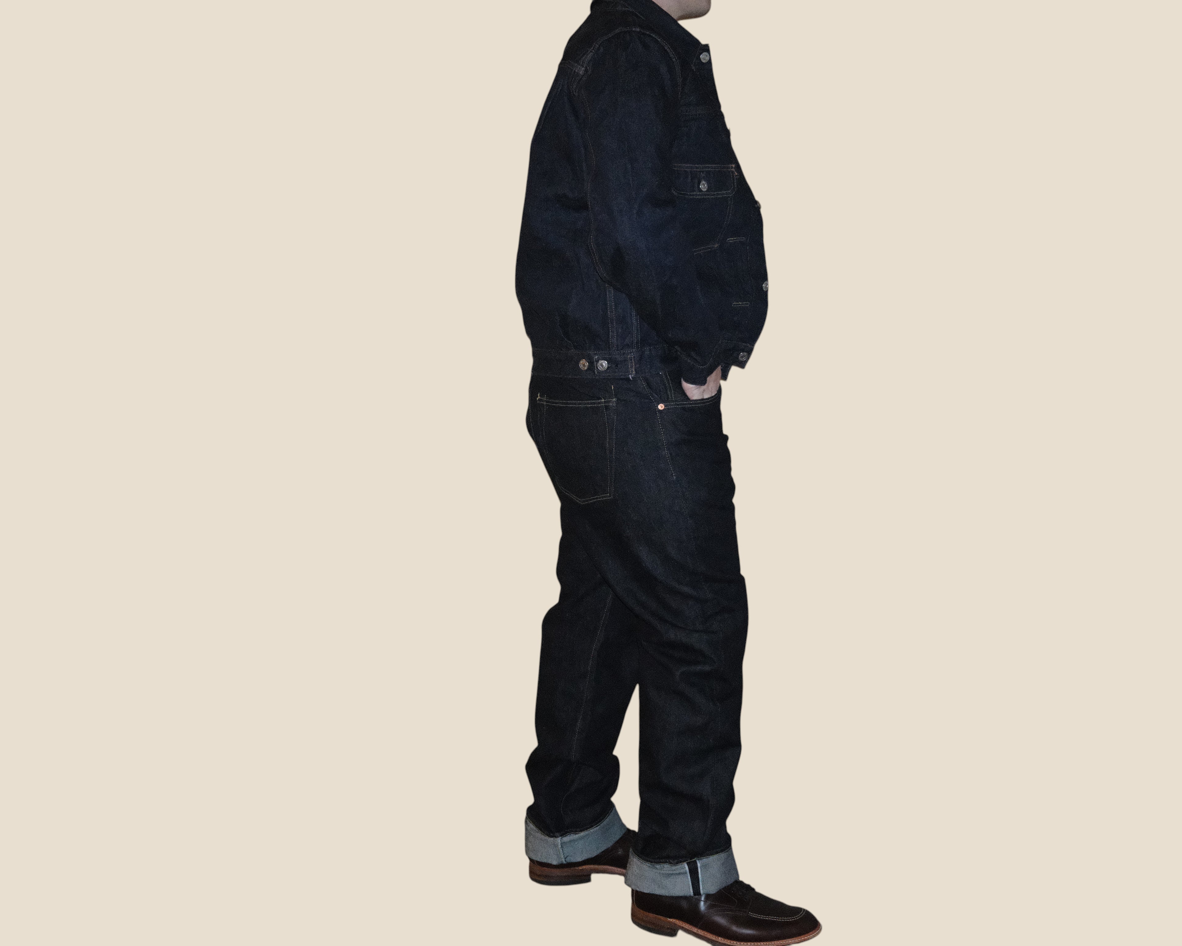 A person shown in side profile wearing dark indigo denim workwear, including a jacket and matching pants with cuffed hems, paired with brown boots, against a plain beige background.