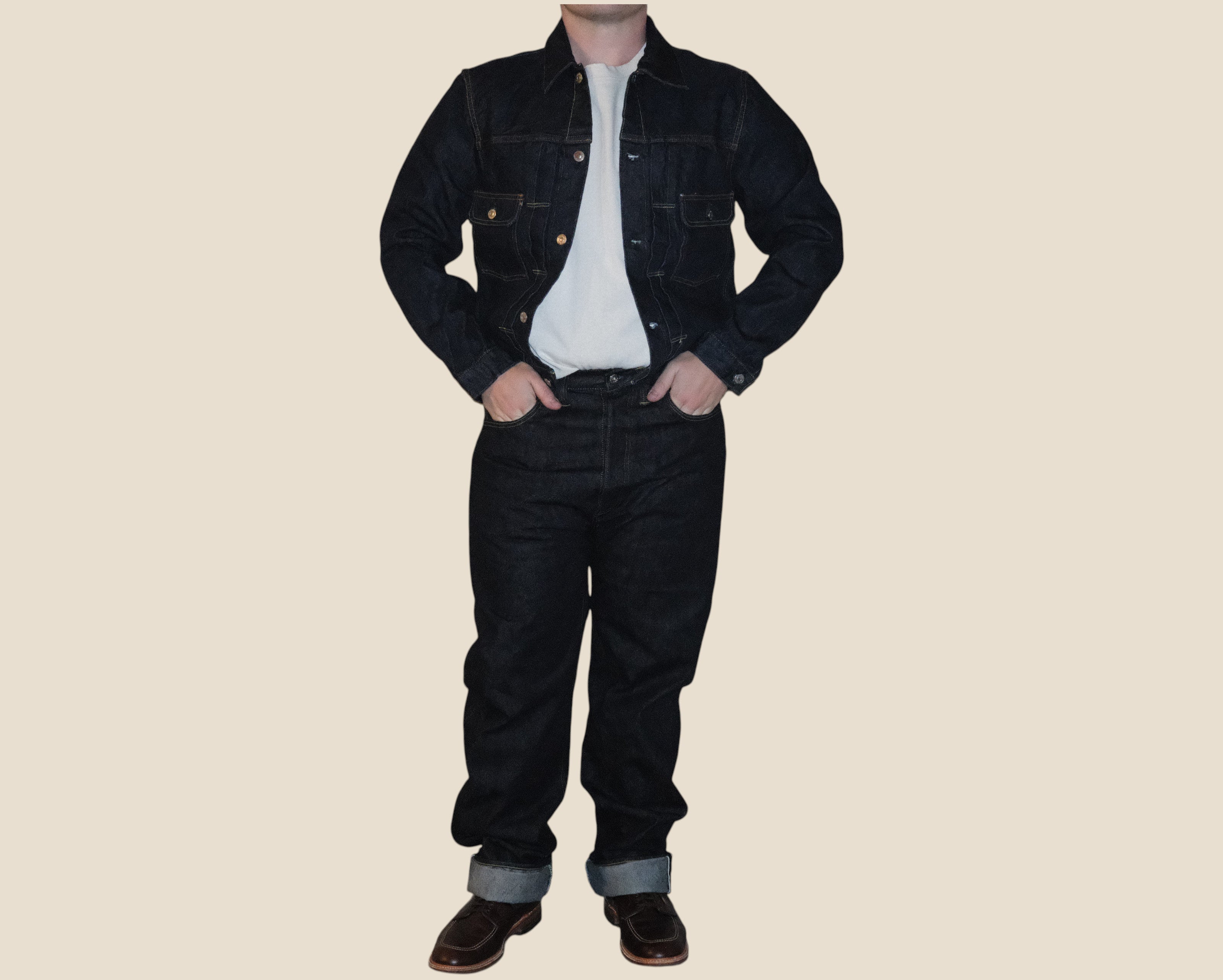 A person wearing a dark denim jacket over a white shirt, dark denim jeans, and brown shoes, standing against a plain beige background.