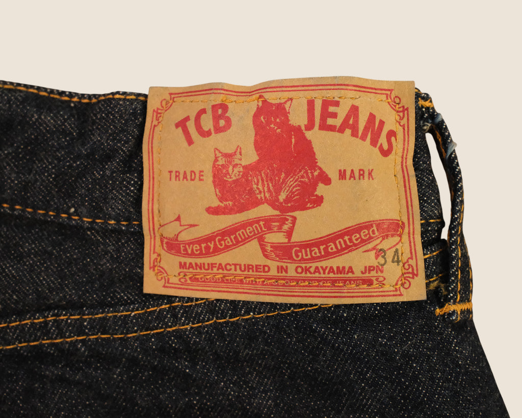 Close-up of a vintage-style denim label on dark blue jeans, featuring red text reading 'TCB JEANS' and 'Trade Mark' with a red cat illustration on a tan background.