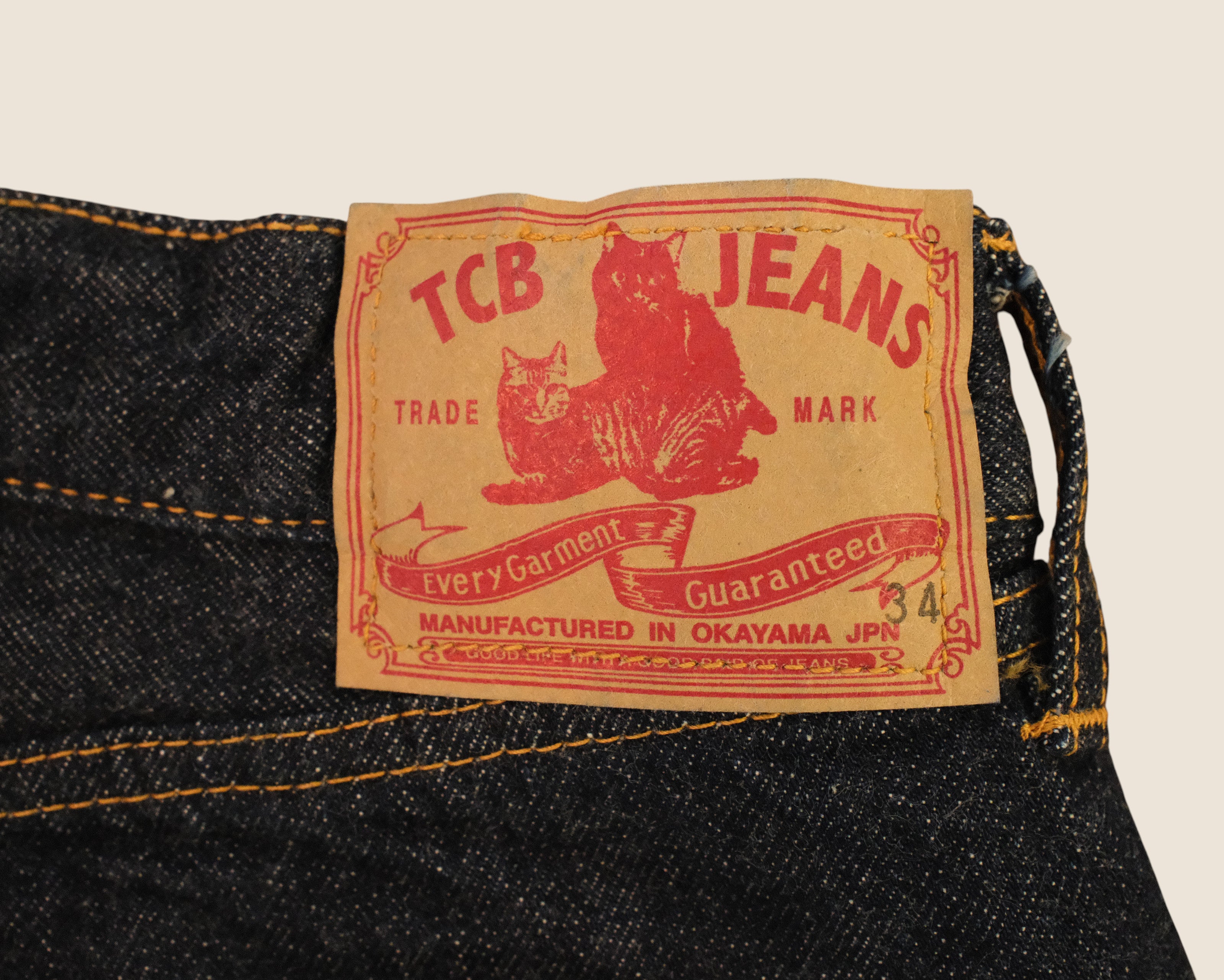 Close-up of a vintage-style denim label on dark blue jeans, featuring red text reading 'TCB JEANS' and 'Trade Mark' with a red cat illustration on a tan background.