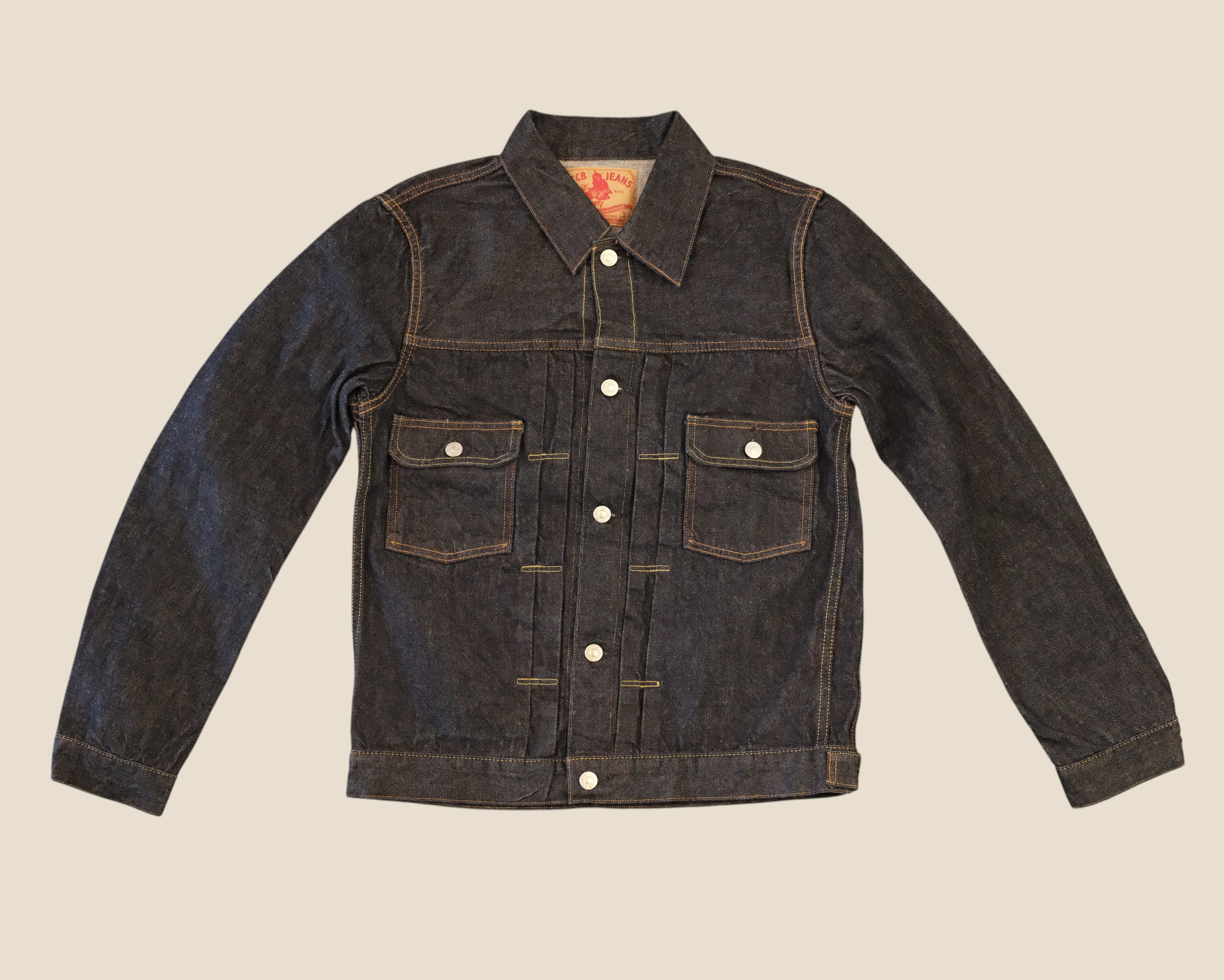 A dark indigo denim jacket with contrast tan stitching, featuring a pointed collar, button-up front, and two buttoned chest pockets, laid flat against a solid beige background.