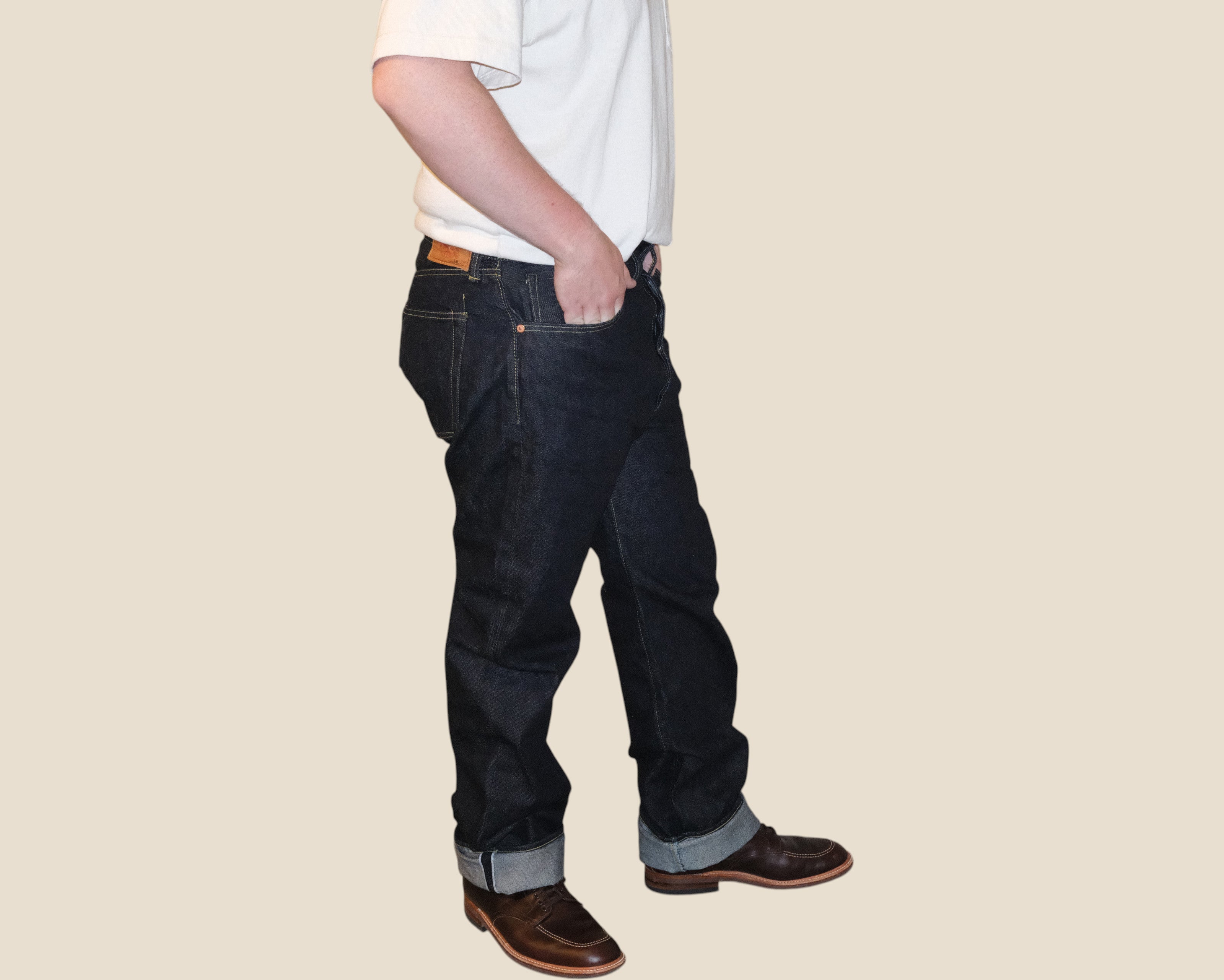 A person shown from the waist down wearing dark blue denim jeans, a white t-shirt, and brown leather shoes, standing against a solid beige background.