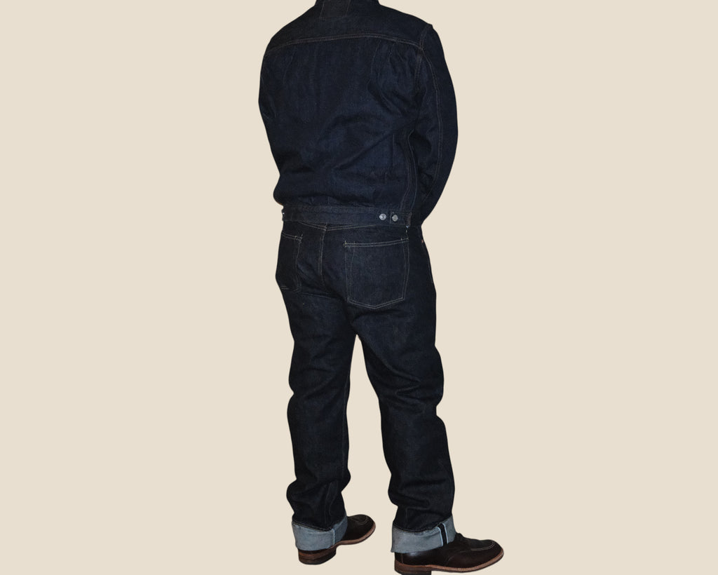A person shown from the back wearing dark indigo denim jeans and a matching dark denim jacket, standing against a solid light beige background.