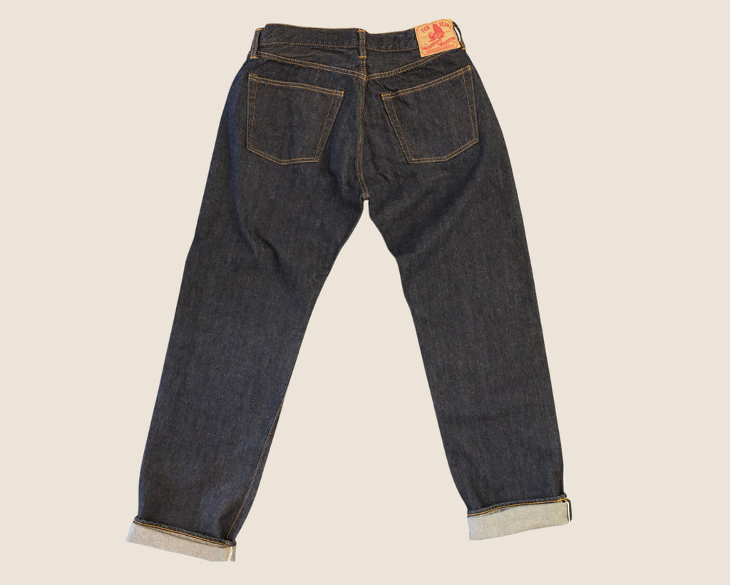 Back view of dark indigo denim jeans with rolled cuffs, showing the back pockets and a small orange brand patch on the waistband, set against a solid light beige background.