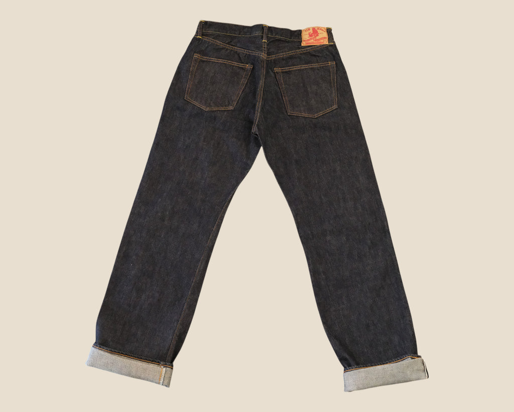 Rear view of dark indigo denim jeans with rolled cuffs, displayed against a solid light beige background.