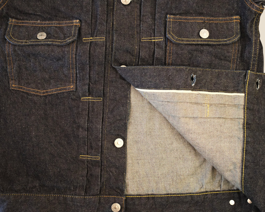 Close-up of a dark indigo denim jacket showing the front pockets, button closure, and a folded pocket revealing a lighter gray inner lining.