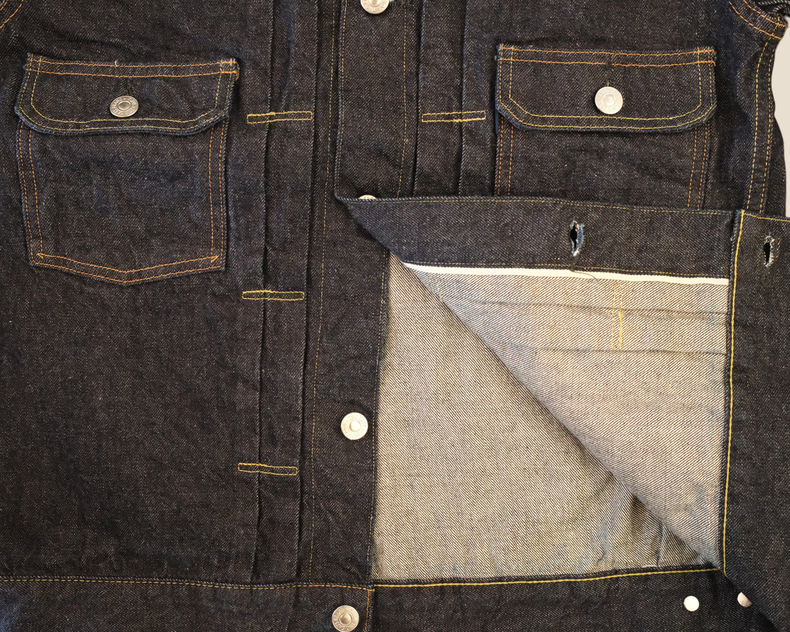 Close-up of a dark indigo denim jacket showing the front pockets, button closure, and a folded pocket revealing a lighter gray inner lining.