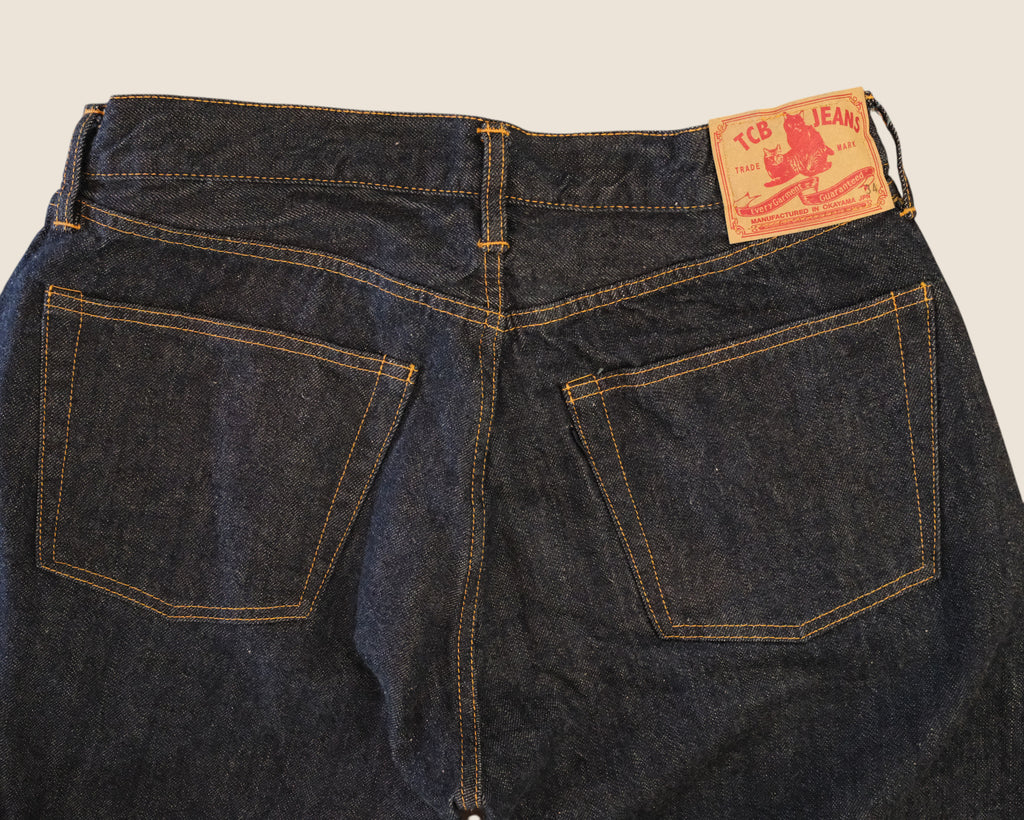 Close-up back view of dark indigo denim jeans showing the waistband, belt loops, and back pockets with contrast stitching. A tan leather patch on the upper right side reads 'ICE JEANS'.