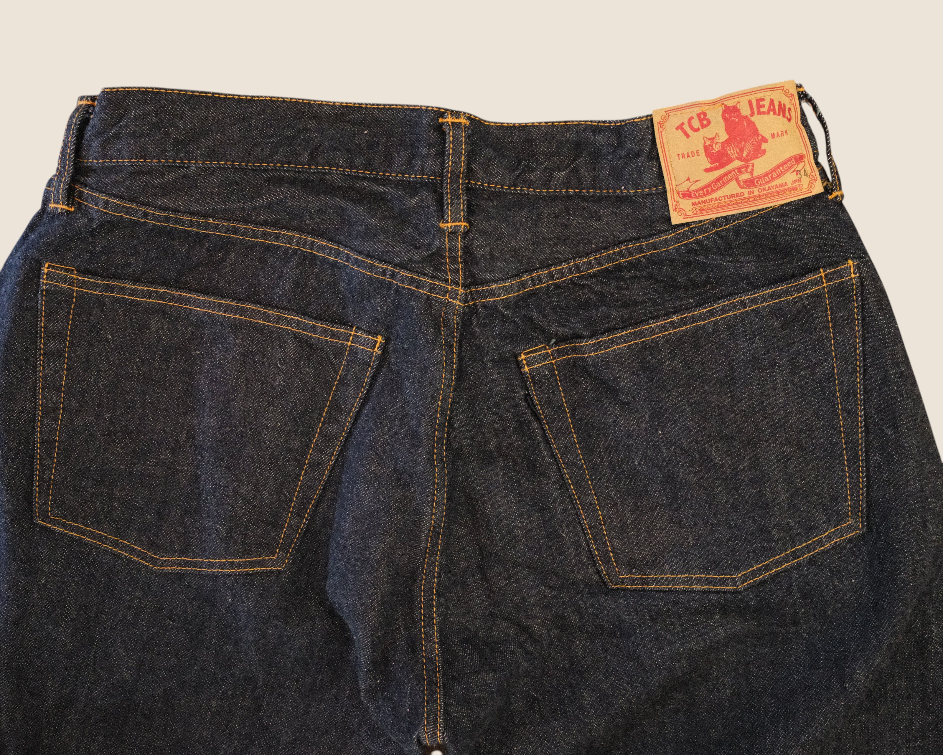 Close-up back view of dark indigo denim jeans showing the waistband, belt loops, and back pockets with contrast stitching. A tan leather patch on the upper right side reads 'ICE JEANS'.