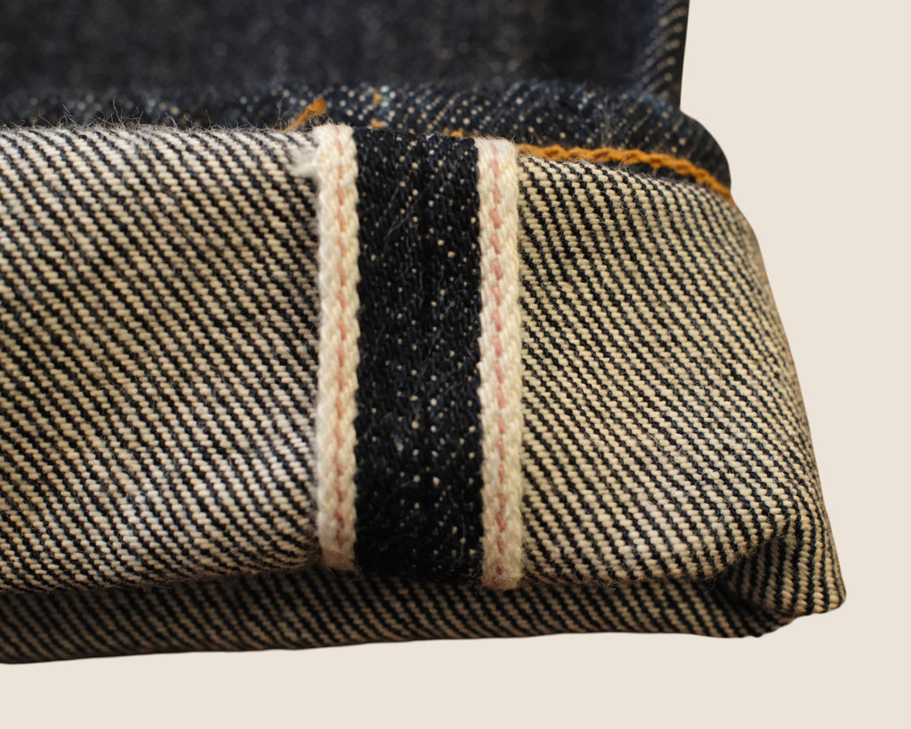 Close-up of folded denim fabric showing a dark blue indigo weave with a central black stripe bordered by two cream-colored contrast stitching lines, and orange-yellow stitching along the top edge.