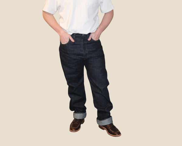 A person wearing dark blue denim jeans with rolled cuffs, a white short-sleeve shirt, and brown shoes, standing against a solid beige background.