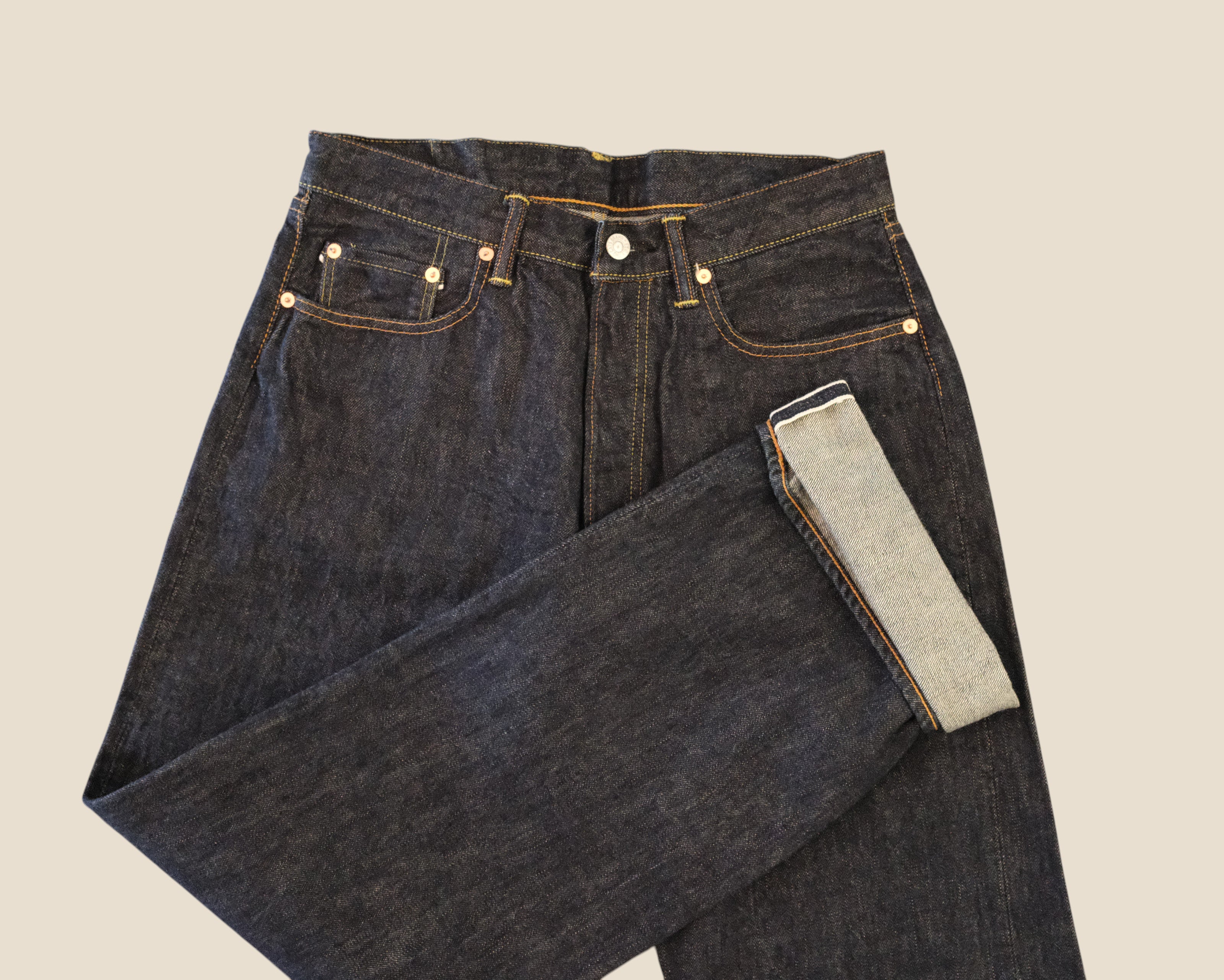 Dark indigo denim jeans folded to show the inner light-wash lining, set against a solid beige background.