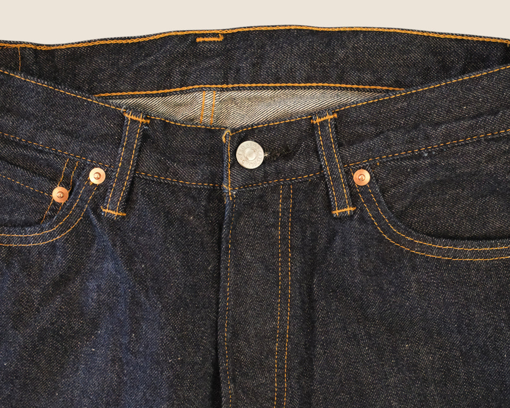 Close-up of dark indigo denim jeans showing the waistband, belt loops, button closure, front pockets, and contrast orange-yellow stitching.