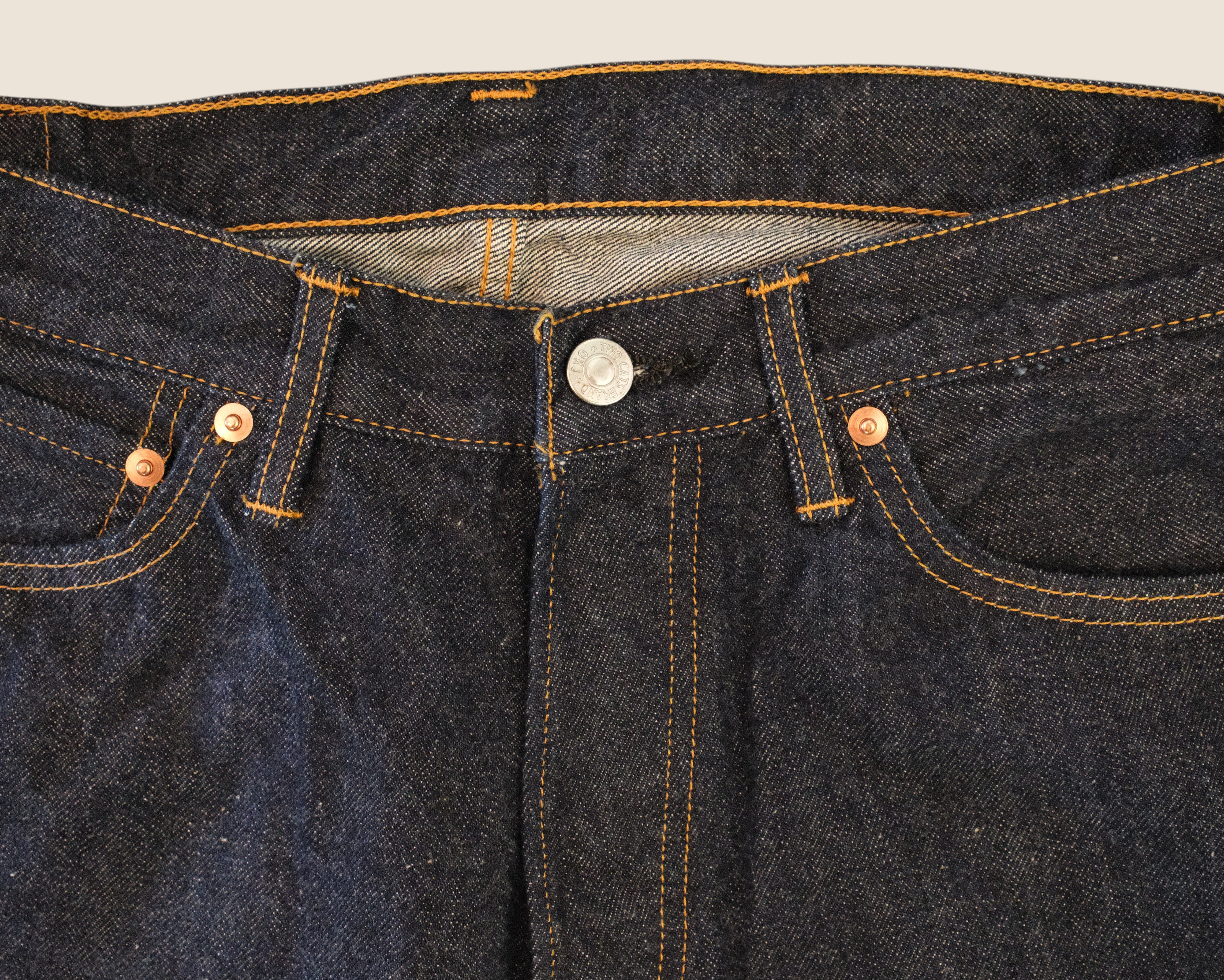Close-up of dark indigo denim jeans showing the waistband, belt loops, button closure, front pockets, and contrast orange-yellow stitching.