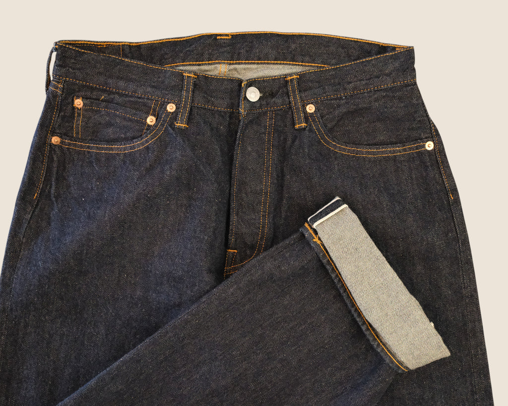 Close-up of dark indigo denim jeans folded to show the inner leg lining and front waistband details against a light beige background.