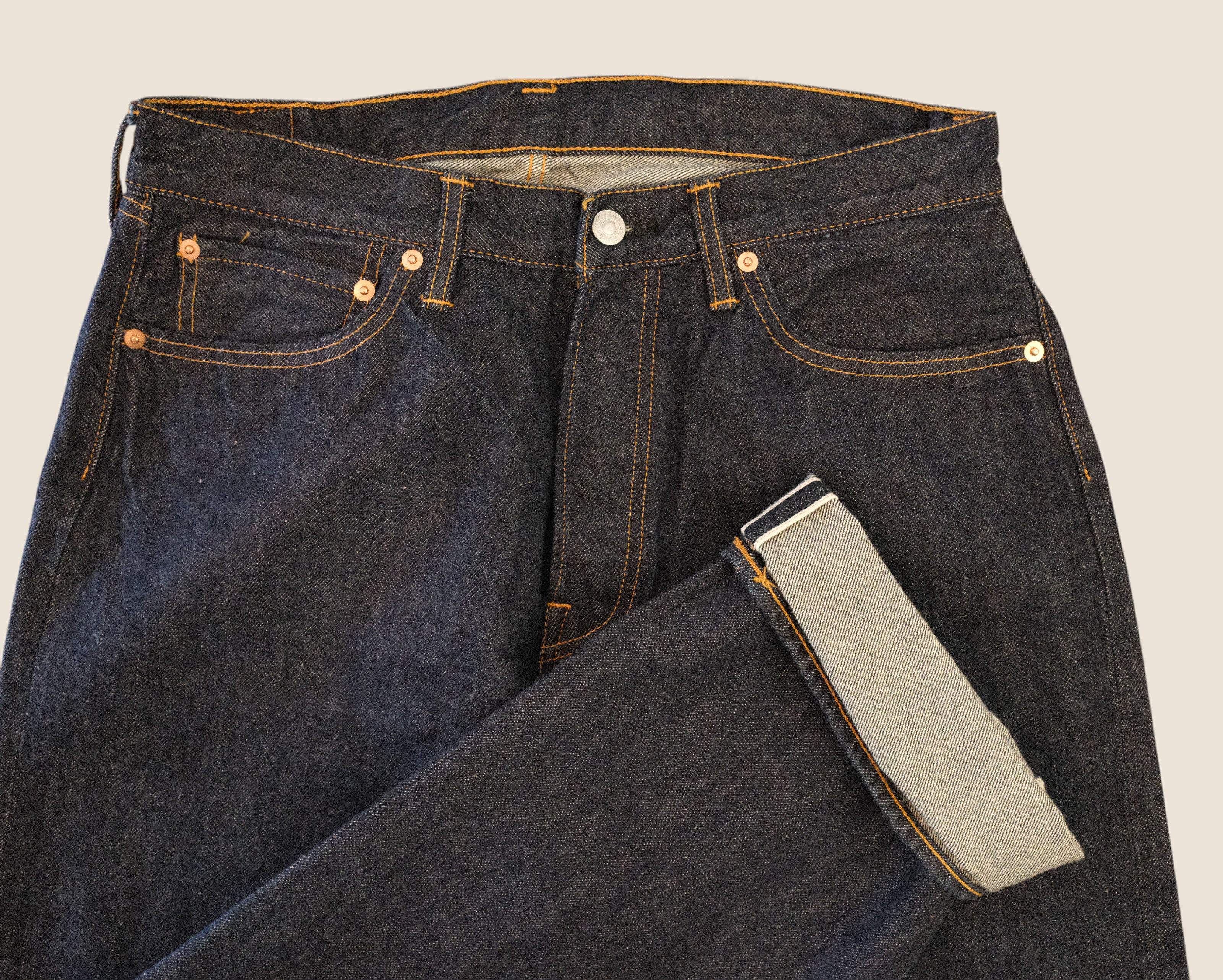 Close-up of dark indigo denim jeans folded to show the inner leg lining and front waistband details against a light beige background.
