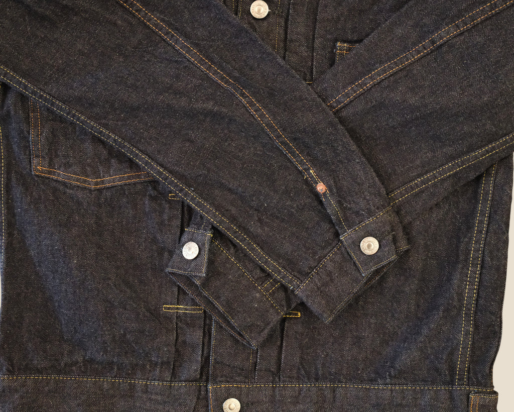 Close-up of a dark indigo denim jacket with contrast tan stitching and silver metal snap buttons, laid flat against a light background.