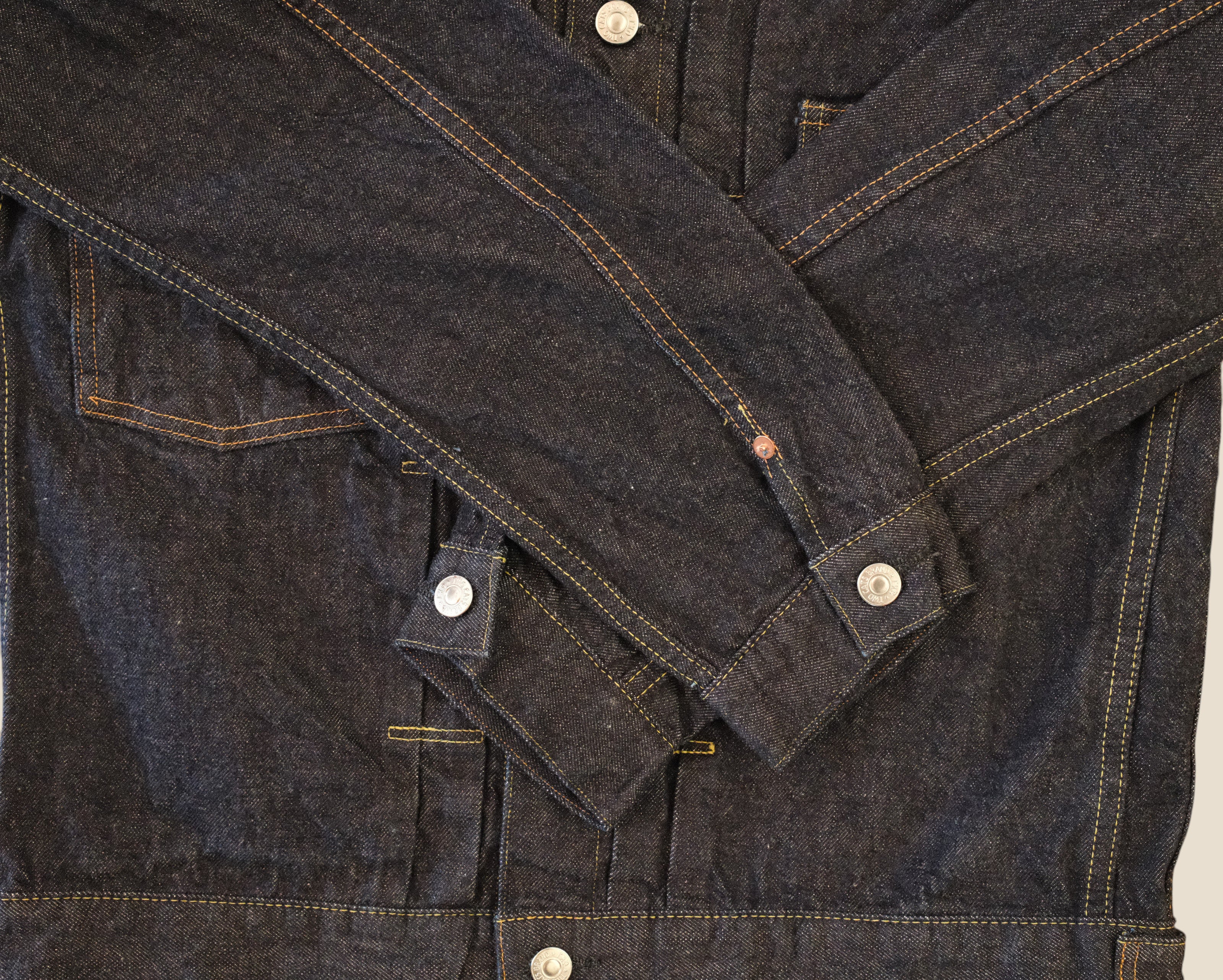 Close-up of a dark indigo denim jacket with contrast tan stitching and silver metal snap buttons, laid flat against a light background.