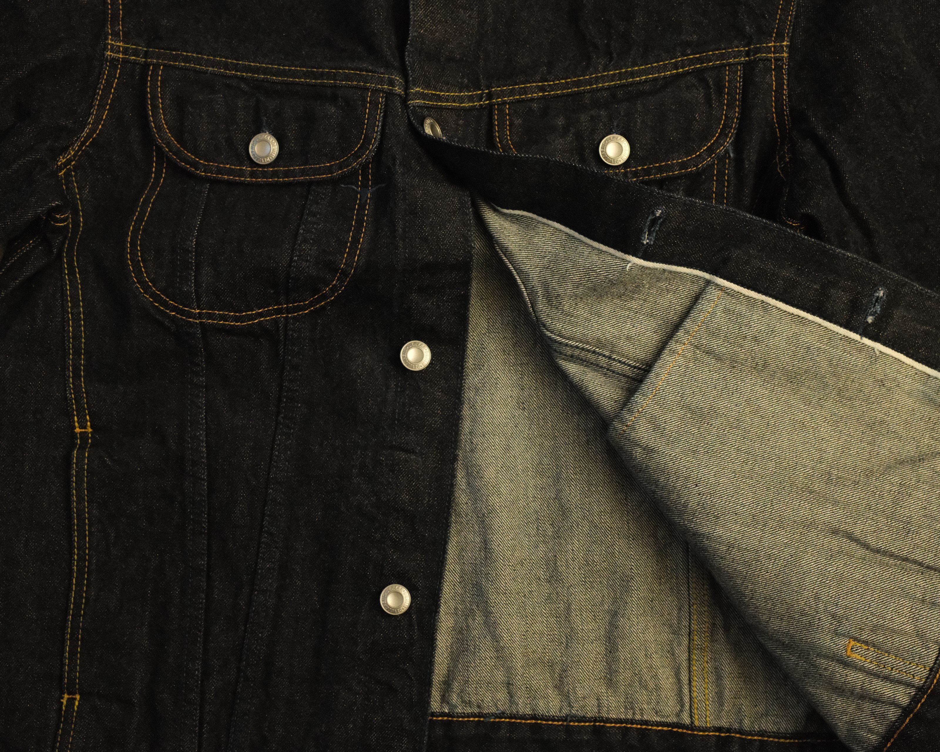 Close-up of a dark indigo denim jacket with contrast yellow stitching, silver snap buttons, and a lighter gray denim lining shown by an open flap pocket.