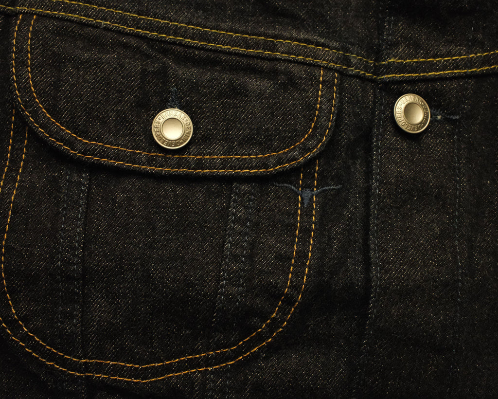 Close-up of dark indigo denim fabric with contrast yellow stitching and two gold-tone metal button snaps on a pocket.
