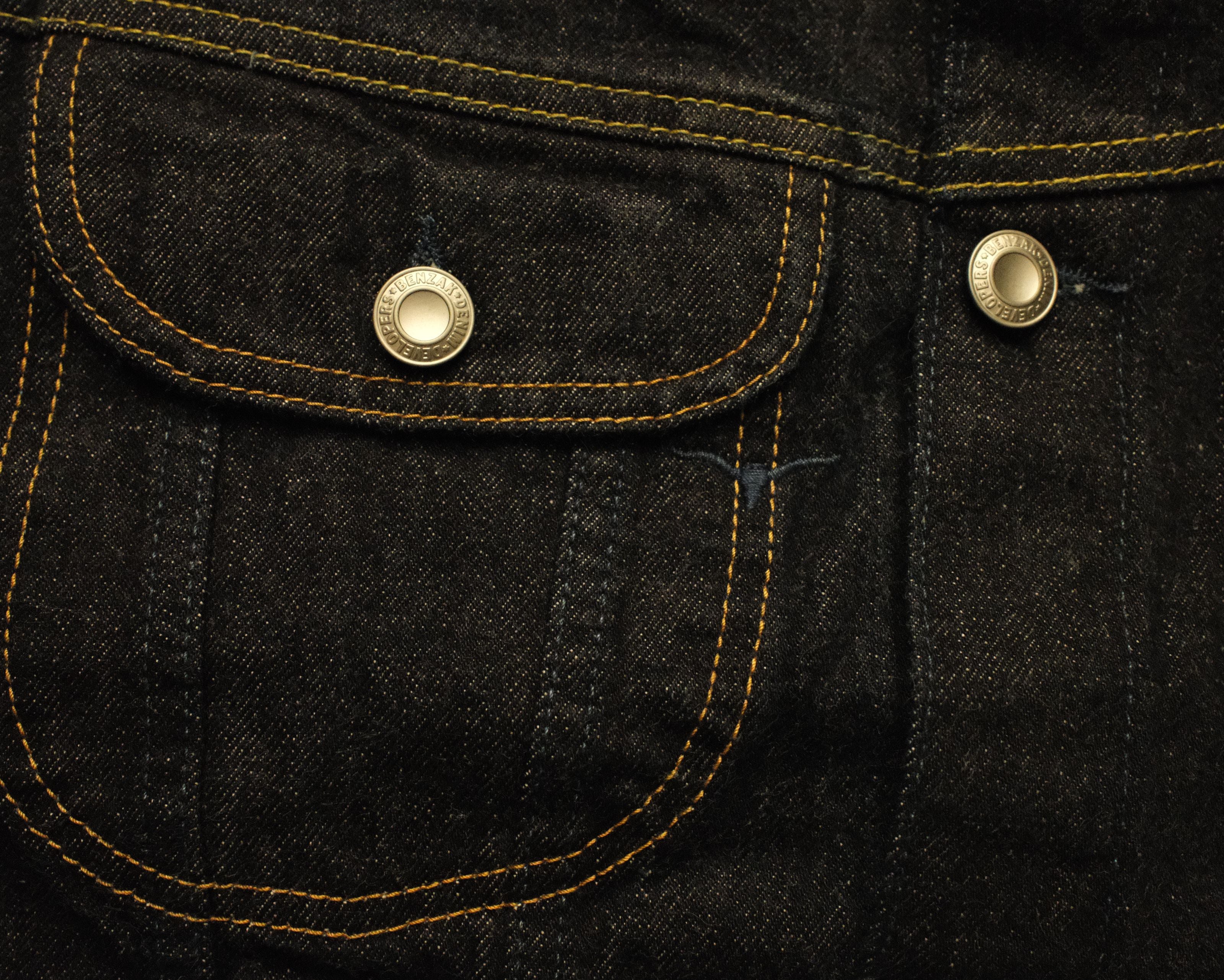 Close-up of dark indigo denim fabric with contrast yellow stitching and two gold-tone metal button snaps on a pocket.